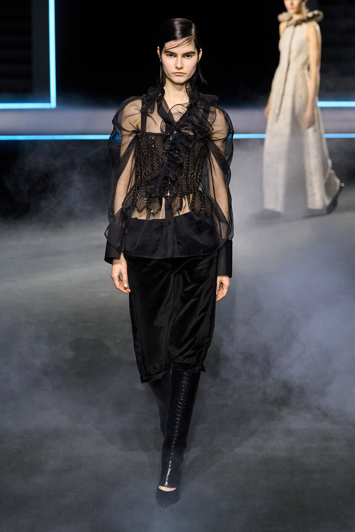 Model walking a black sheer blouse with a beaded corset-like bodice and voluminous sleeves, paired with black pants and knee-high boots on a smoky runway.