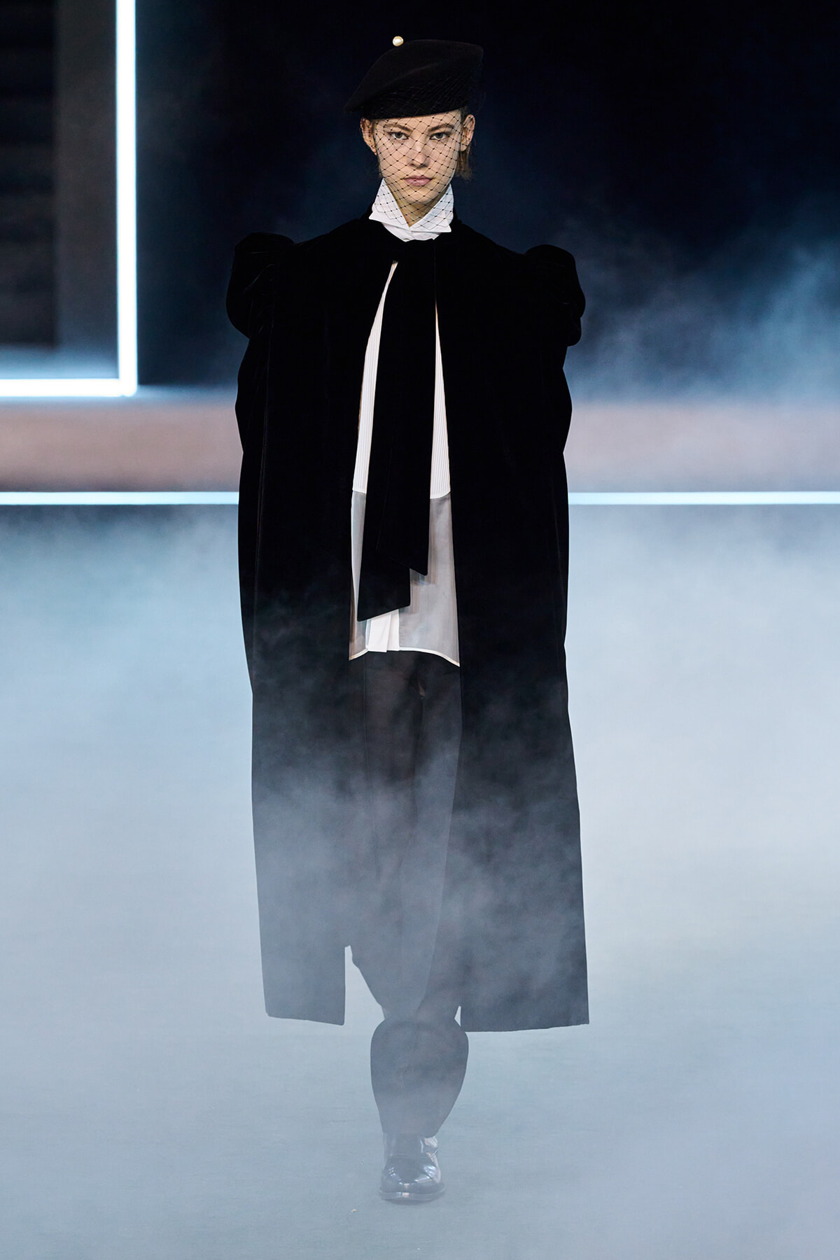 Model walks a smoky runway in a black cape, white blouse, wide belt-like black bow, and a black hat with netted veil.