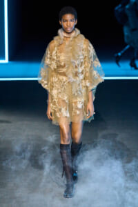 Model walks a fashion runway wearing a sheer beige lace dress with ruffles, knee-high socks, and black boots.