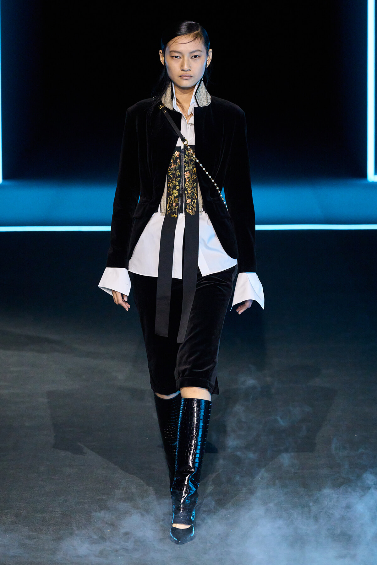 Model on a runway wearing a black velvet blazer, white shirt, ornate tie-front, and shiny knee-high boots against a blue neon-lit stage.