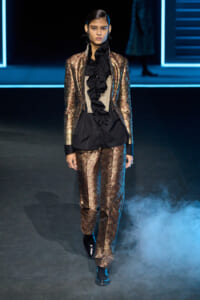 Model walks a runway in a bronze metallic brocade suit with black ruffled blouse and beige corset, black boots.
