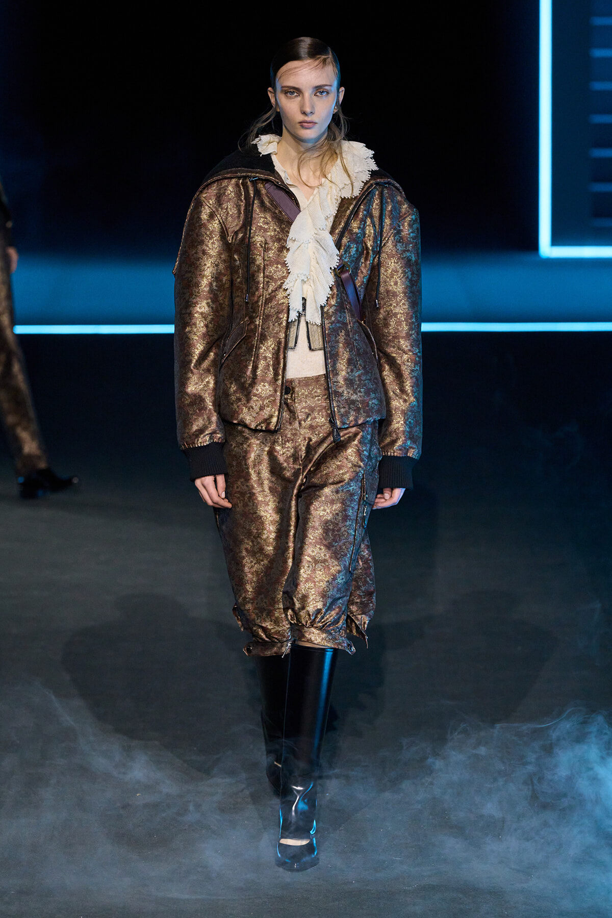Model walking a runway in a bronze metallic suit with a white ruffled blouse and black knee-high boots, on a smoky catwalk.] "]} (Note: Oops)