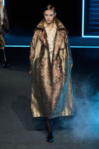 Model walking the runway in a long metallic bronze-gold brocade trench coat over a cream ruffled blouse.