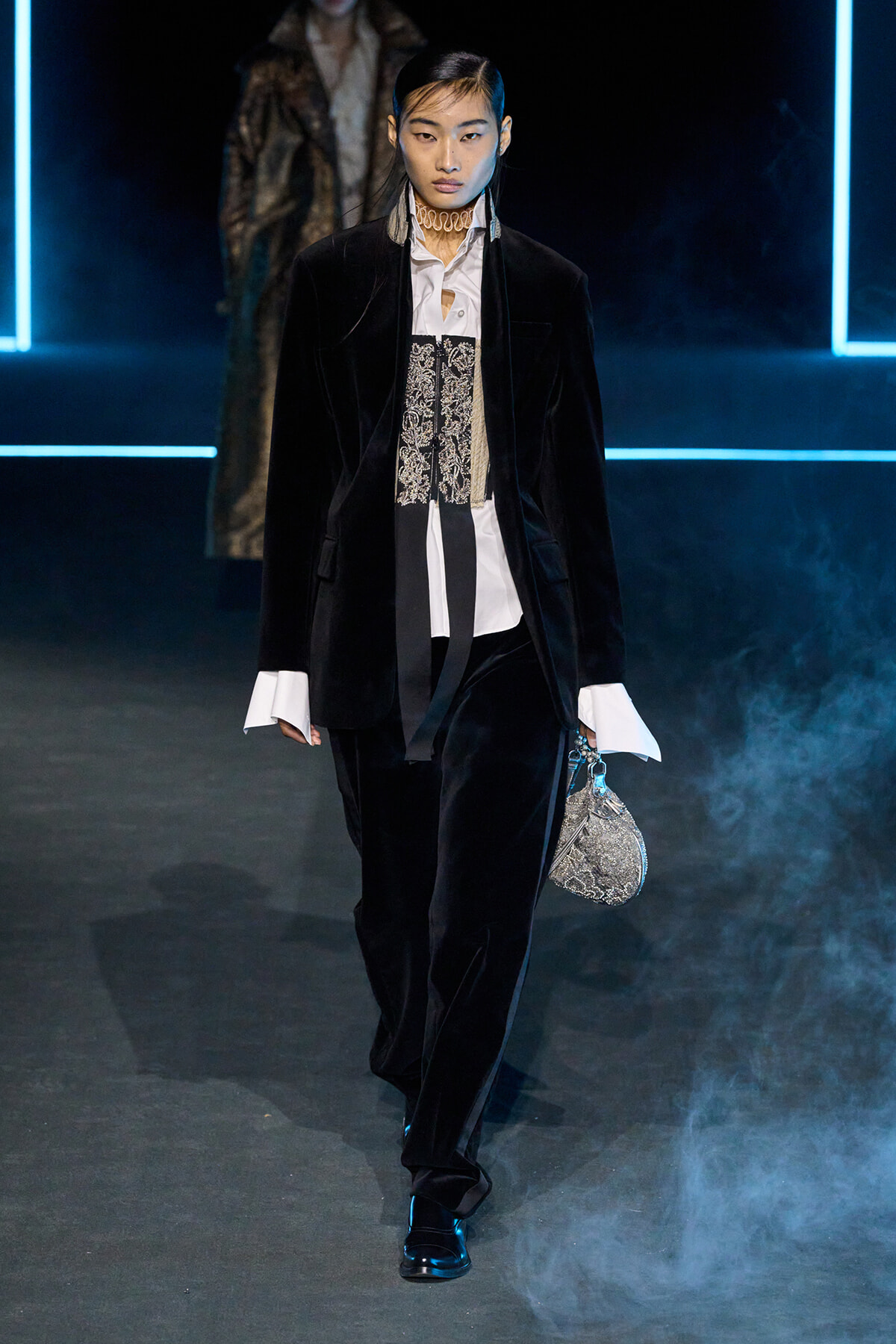 Model walking a fashion runway in a black velvet suit with a white shirt, lace corset detailing, and a small metallic sequined bag.