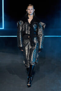 Model wearing a metallic silver oversized jacket and matching pants with a black scarf on a blue-lit fashion runway.