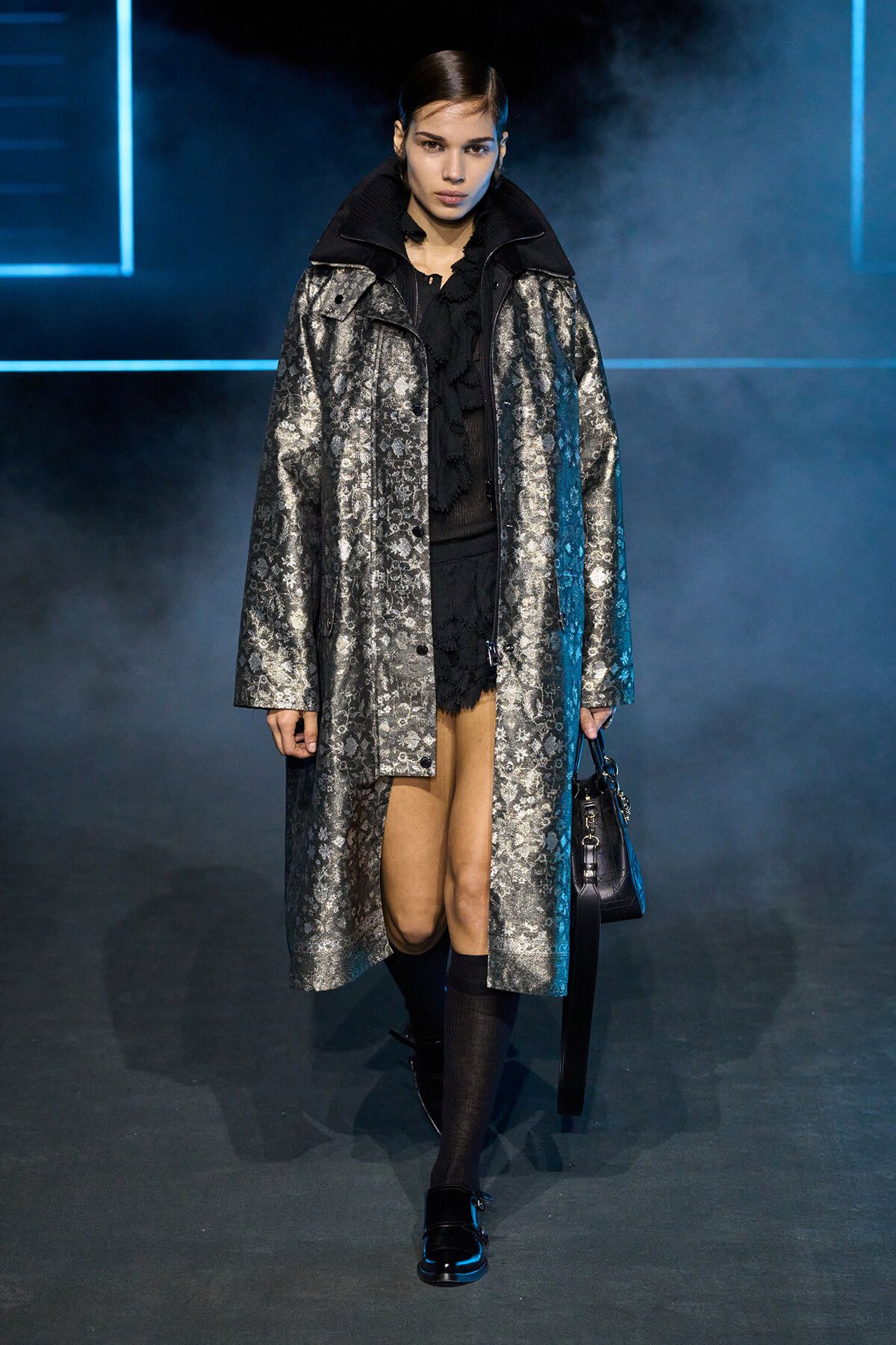 Model walking runway in a shiny silver patterned coat over black outfit, holding a black handbag.