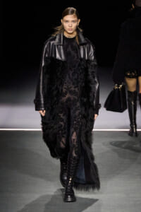 Model on a runway wearing a black fur-trimmed coat over a sheer black lace jumpsuit, with lace-up boots and a chunky gold choker.