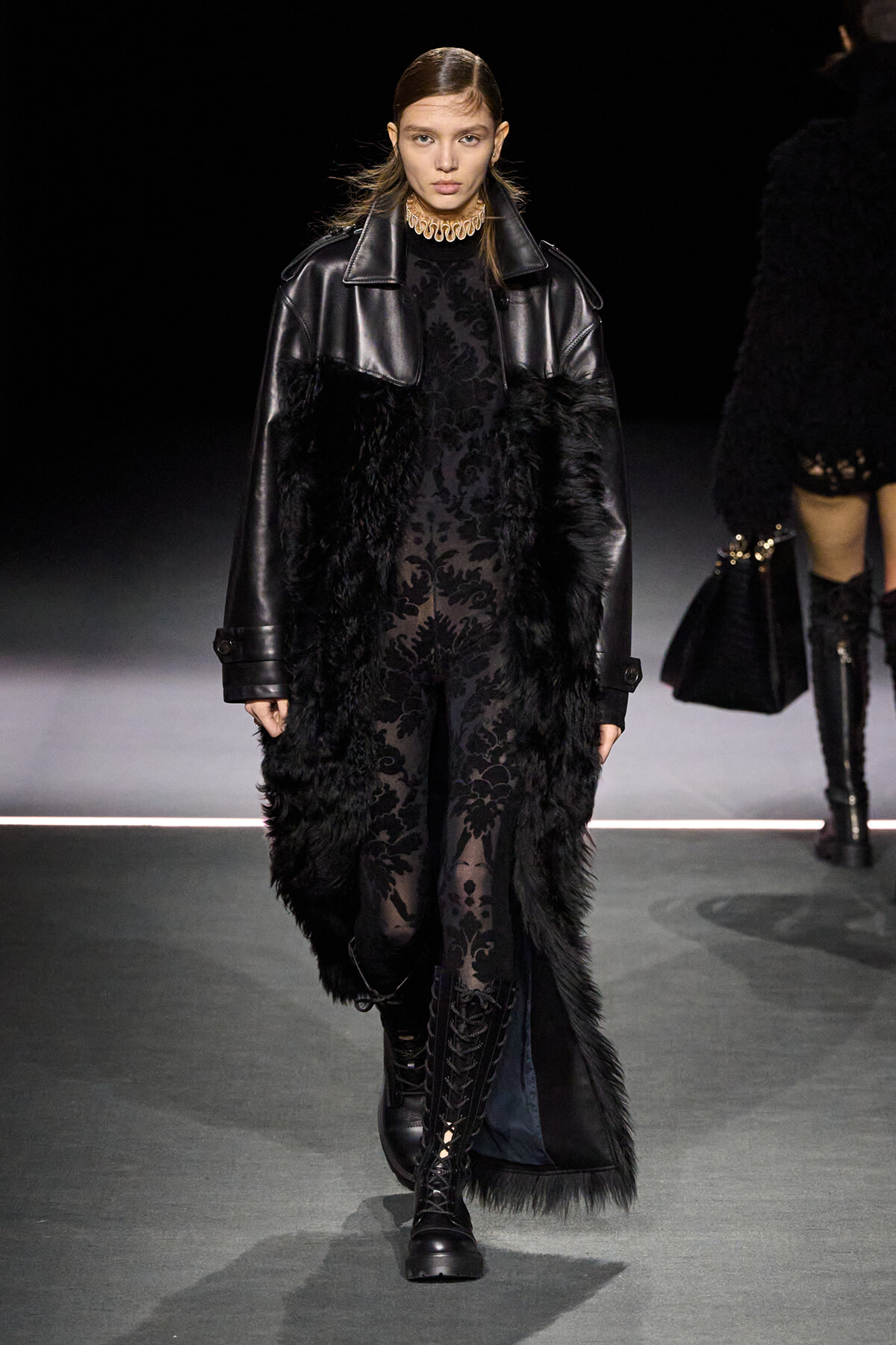 Model on a runway wearing a black fur-trimmed coat over a sheer black lace jumpsuit, with lace-up boots and a chunky gold choker.