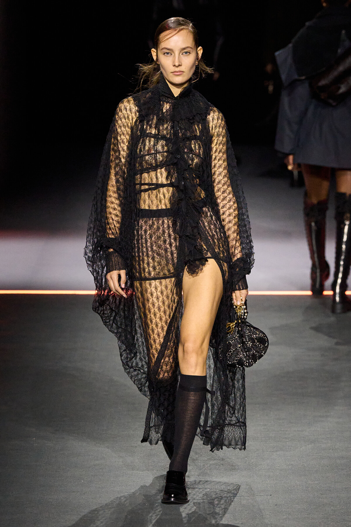 Runway model in a sheer black lace gown with a matching cape, carrying a small black beaded handbag.