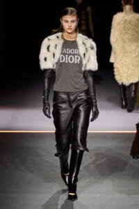 Model walking the runway in a white fur shrug over a gray DIOR tee, with black leather pants, gloves, and knee-high boots.