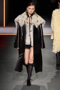 Model on a runway wearing a black leather coat with a white fur collar, black lace shorts, knee-high socks, and black shoes.
