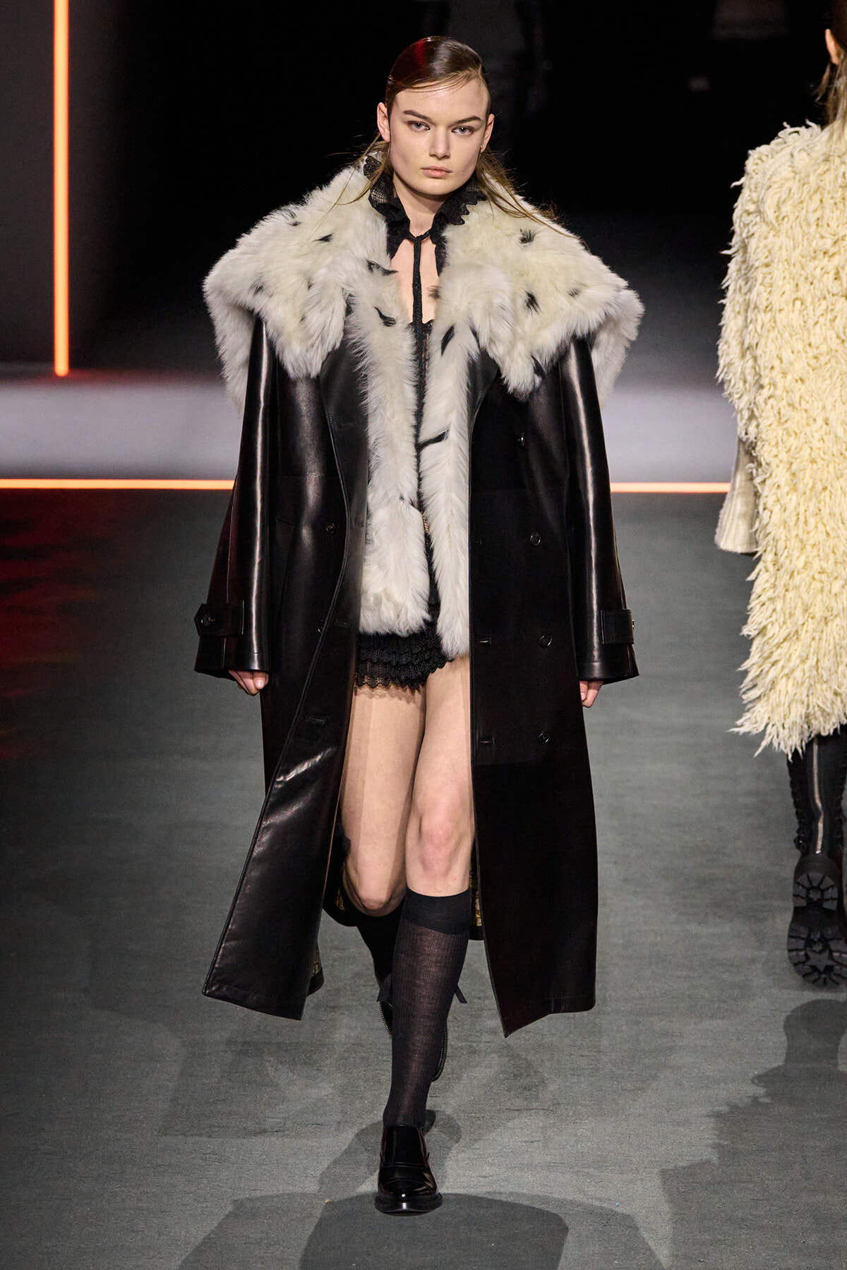 Model on a runway wearing a black leather coat with a white fur collar, black lace shorts, knee-high socks, and black shoes.