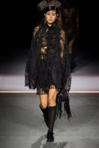 Runway model in a sheer black lace dress with cape, walking the catwalk with a fringed black bag in hand.