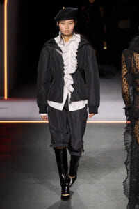 Model walking a fashion runway in a black jacket over a white ruffled blouse, black baggy pants, glossy knee-high boots, and a black beret with net veil.