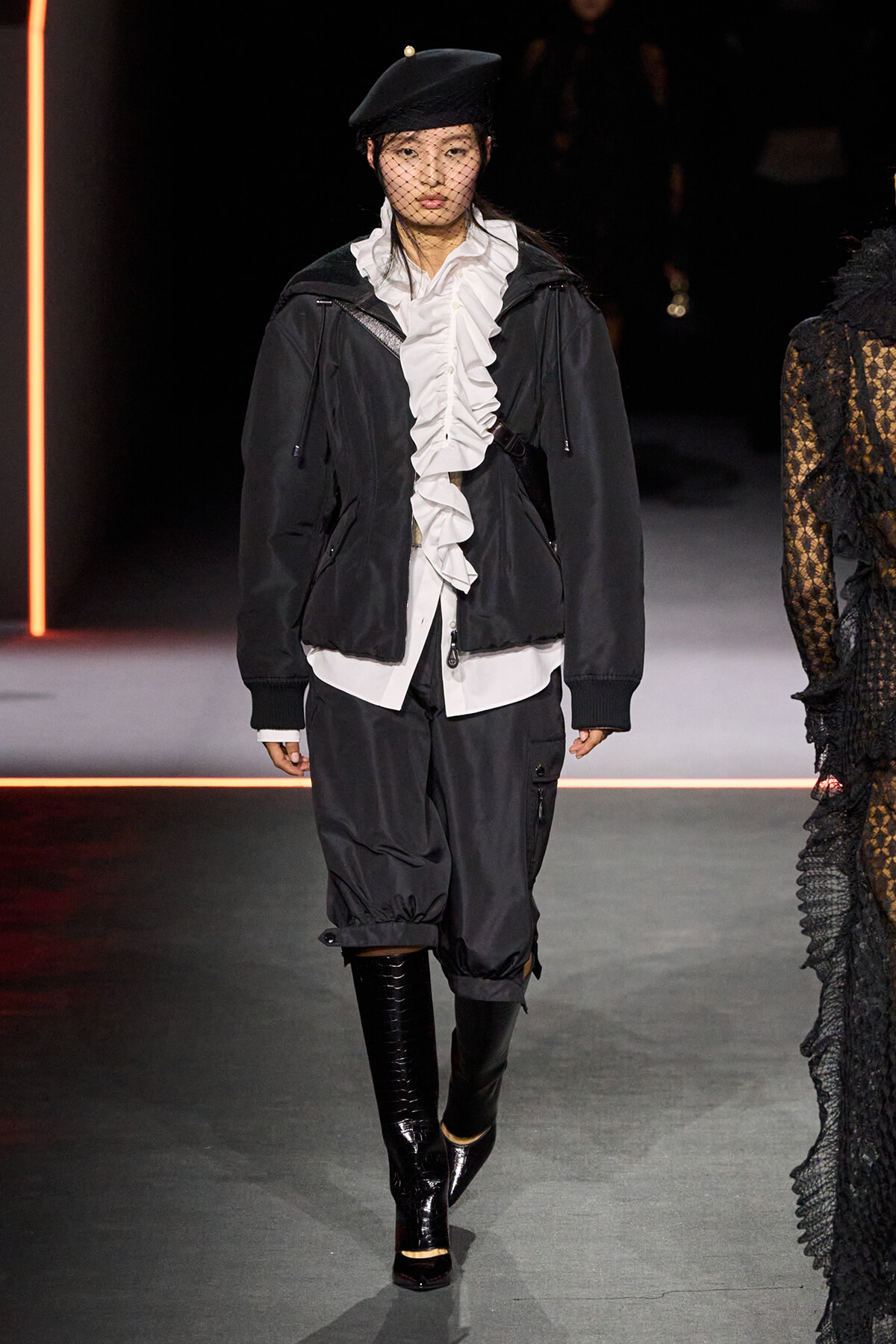 Model walking a fashion runway in a black jacket over a white ruffled blouse, black baggy pants, glossy knee-high boots, and a black beret with net veil.