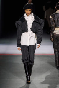 Runway model wearing a black oversized jacket over a white ruffled blouse, black cropped pants, and shiny knee-high boots with a black beret and veil smoothing forward.