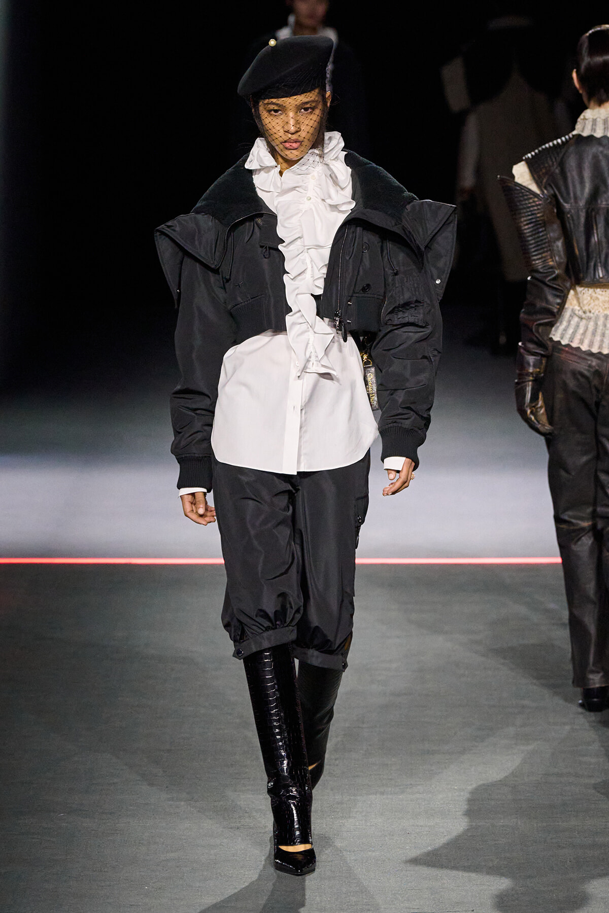 Runway model wearing a black oversized jacket over a white ruffled blouse, black cropped pants, and shiny knee-high boots with a black beret and veil smoothing forward.