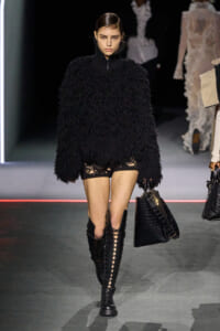 Model walks the runway in an oversized black shaggy sweater, black lace-up knee-high boots, and distressed shorts with fishnet tights, carrying a black handbag.