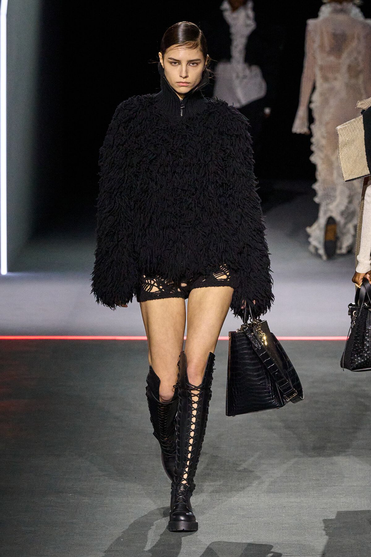 Model walks the runway in an oversized black shaggy sweater, black lace-up knee-high boots, and distressed shorts with fishnet tights, carrying a black handbag.