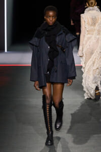 Female model walks a fashion runway in a navy oversized coat, black scarf, and tall lace-up knee-high boots.