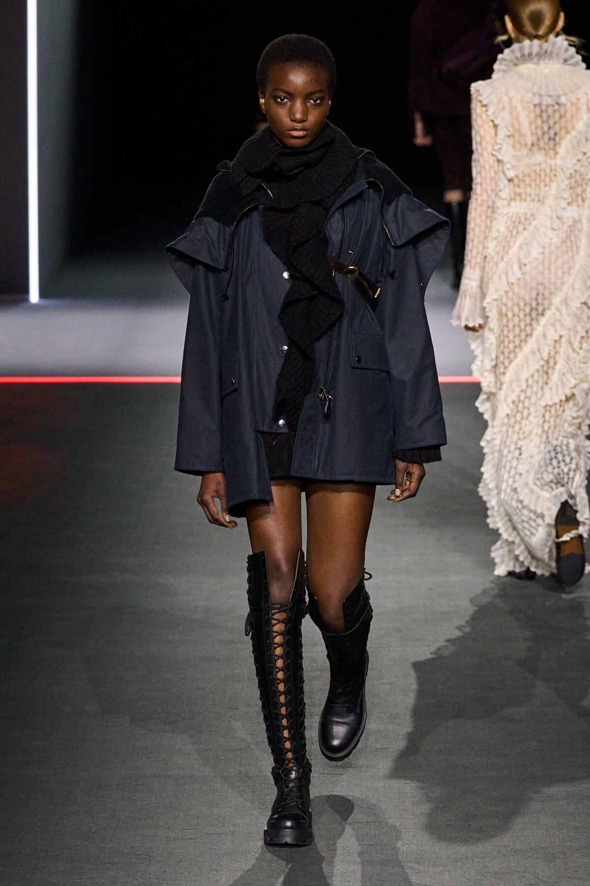 Female model walks a fashion runway in a navy oversized coat, black scarf, and tall lace-up knee-high boots.