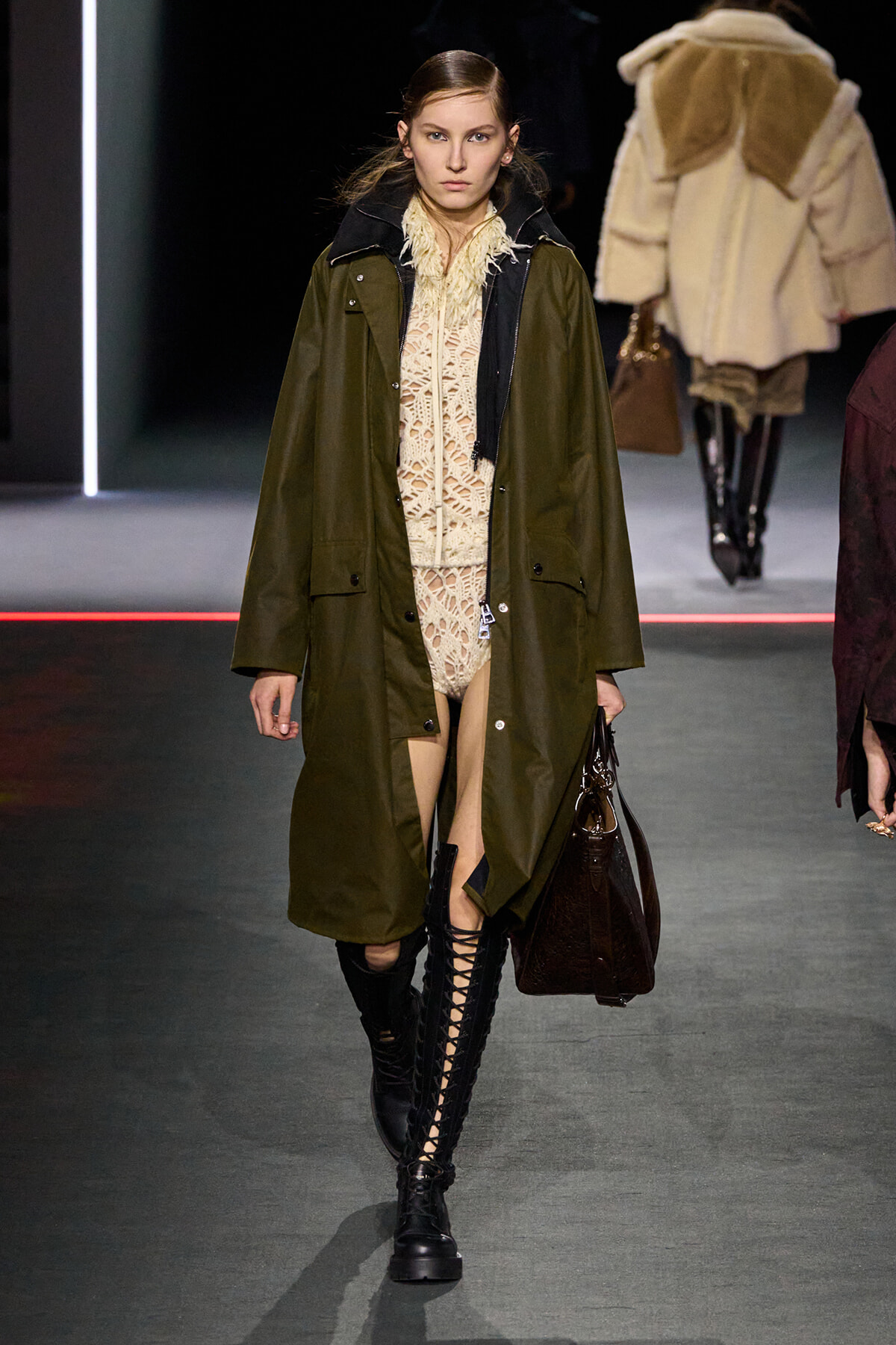 Model walks the runway in an olive-green coat over a beige lace outfit, paired with tall black lace-up boots and a brown handbag.