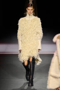 Model walks a fashion show wearing a cream shaggy knit dress with a high zip collar and knee-high black lace-up boots.