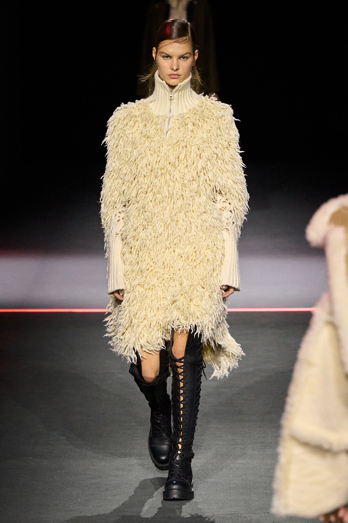 Model walks a fashion show wearing a cream shaggy knit dress with a high zip collar and knee-high black lace-up boots.