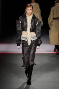 Model walking a fashion show in a black leather layered jacket over a cream lace blouse and matching dark leather skirt with knee-high boots.
