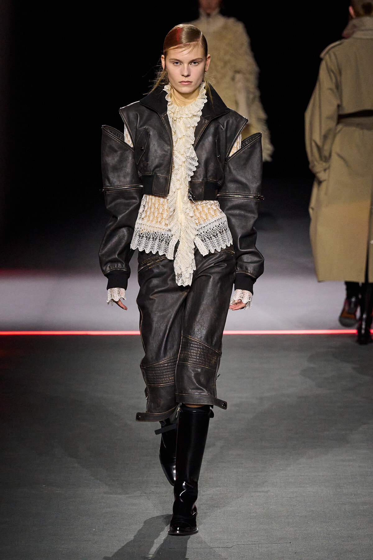 Model walking a fashion show in a black leather layered jacket over a cream lace blouse and matching dark leather skirt with knee-high boots.