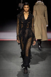 Model walks the runway in a floor-length sheer black lace dress with ruffled trim and a choker necklace.