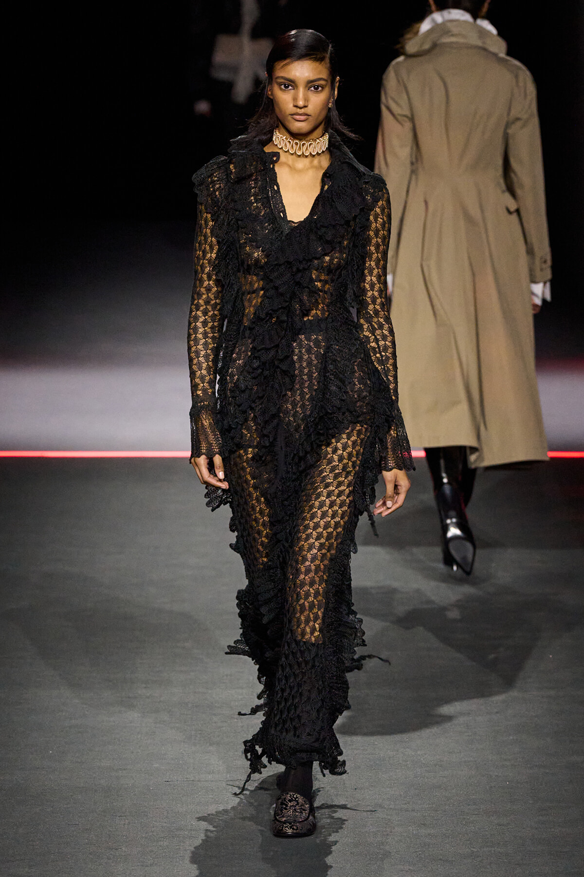 Model walks the runway in a floor-length sheer black lace dress with ruffled trim and a choker necklace.