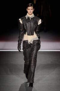 Model walks the runway in a black leather suit with a lace-trimmed cream blouse and structured shoulders.