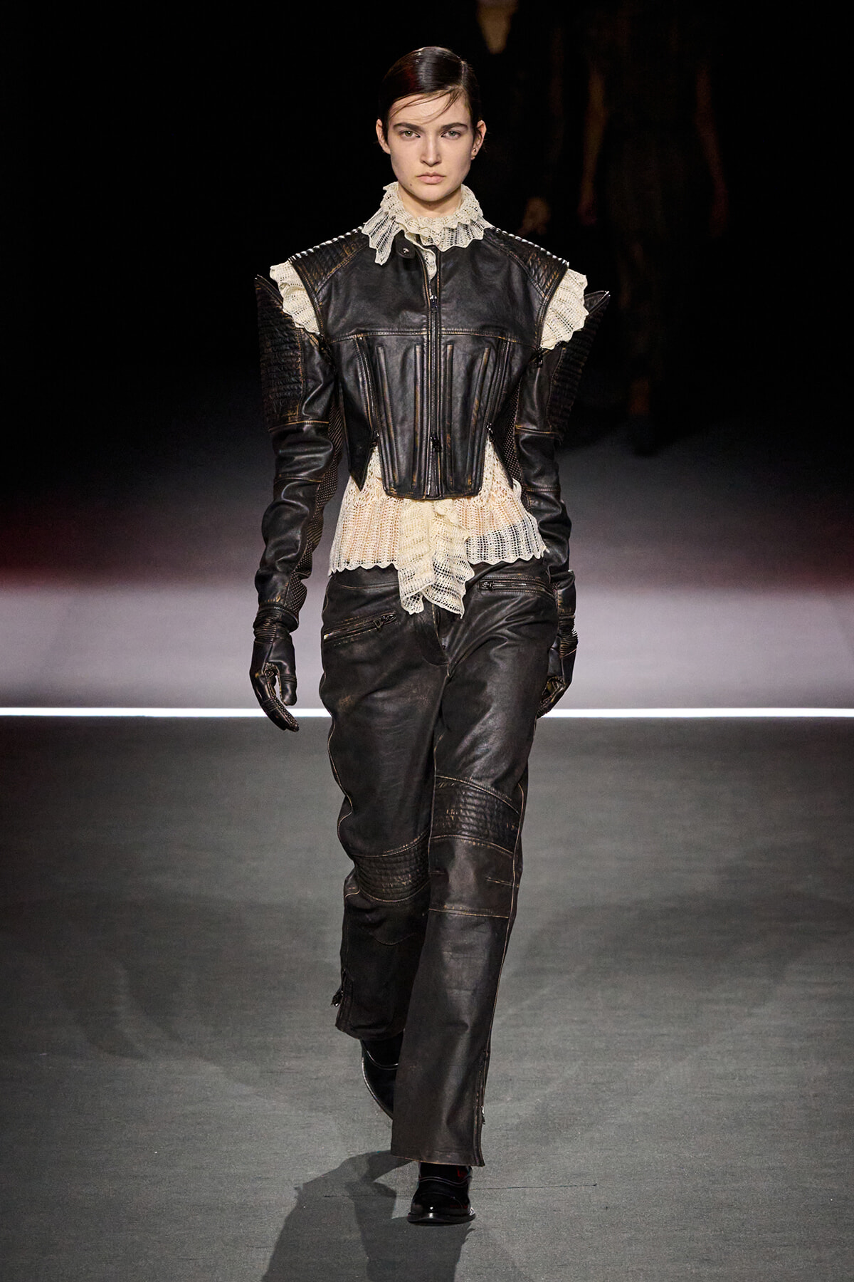 Model walks the runway in a black leather suit with a lace-trimmed cream blouse and structured shoulders.