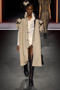 Model walks the runway in a long beige textured coat over a cream ruffled blouse, paired with tall black lace-up boots.