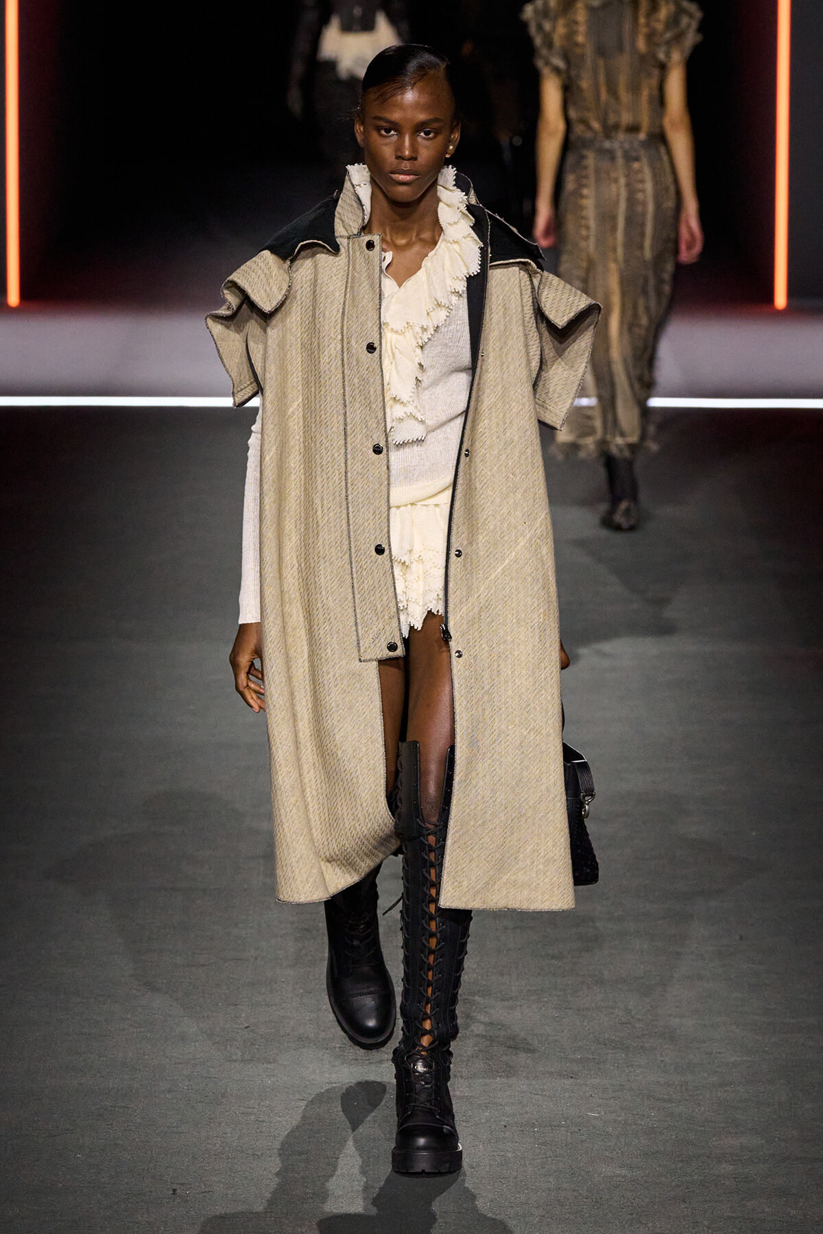 Model walks the runway in a long beige textured coat over a cream ruffled blouse, paired with tall black lace-up boots.