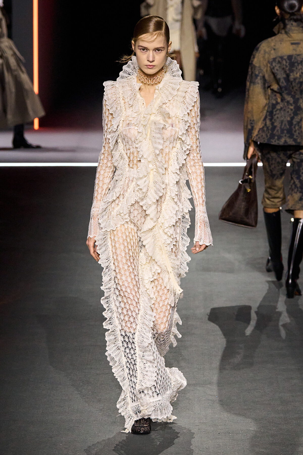 Model walking a runway in an ivory lace gown with cascading ruffles and a high frilled collar.