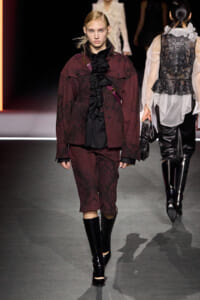 Model on a runway wearing a burgundy patterned suit with a black ruffled blouse, knee-length shorts, and tall black boots.