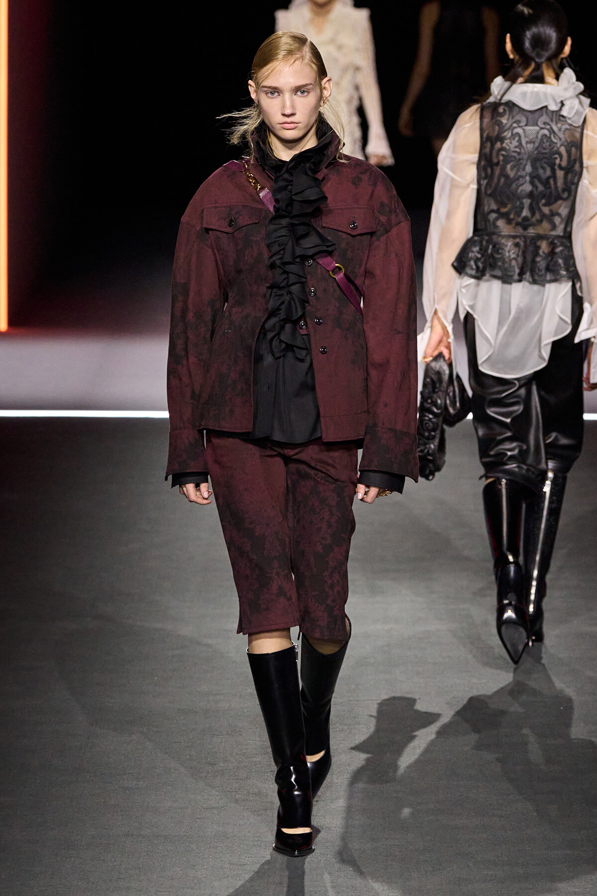 Model on a runway wearing a burgundy patterned suit with a black ruffled blouse, knee-length shorts, and tall black boots.