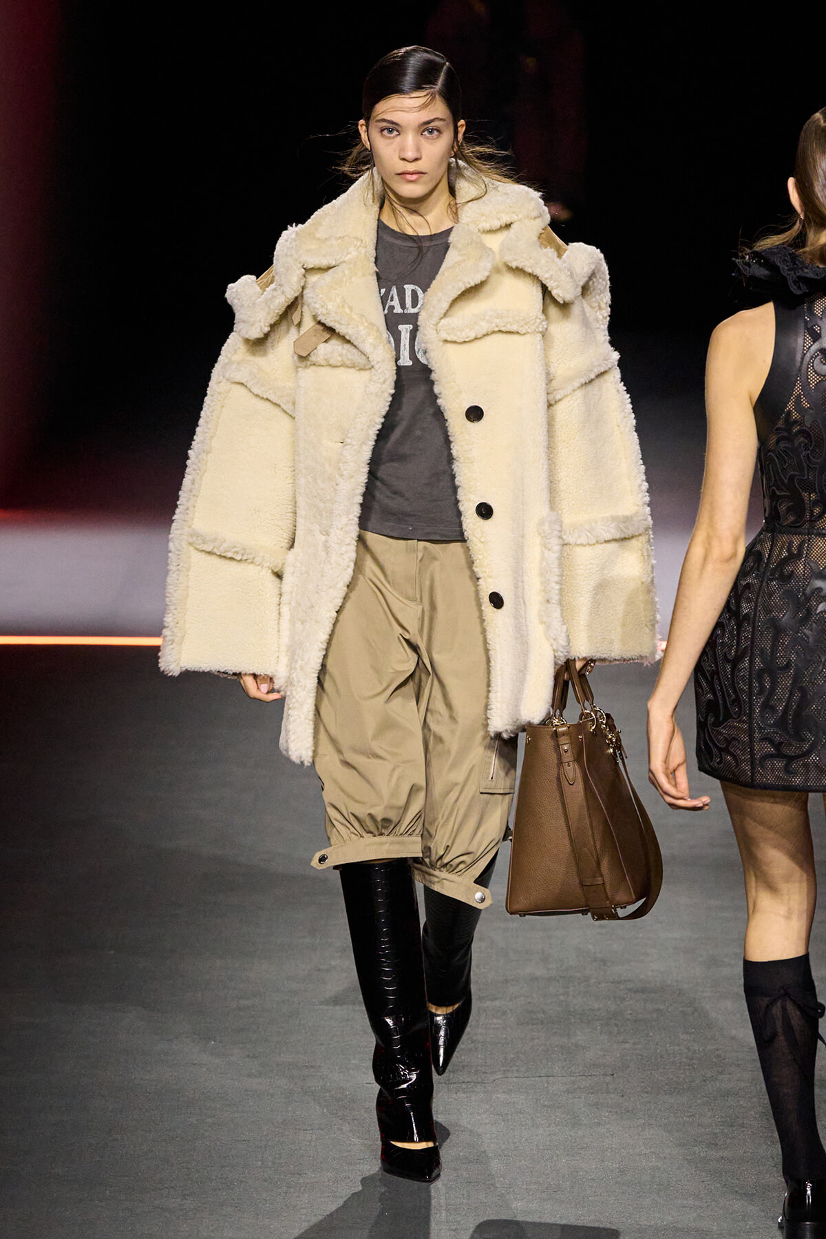 Model on the runway wearing an oversized cream shearling coat, tan cargo pants, black knee-high boots, and carrying a brown structured handbag.