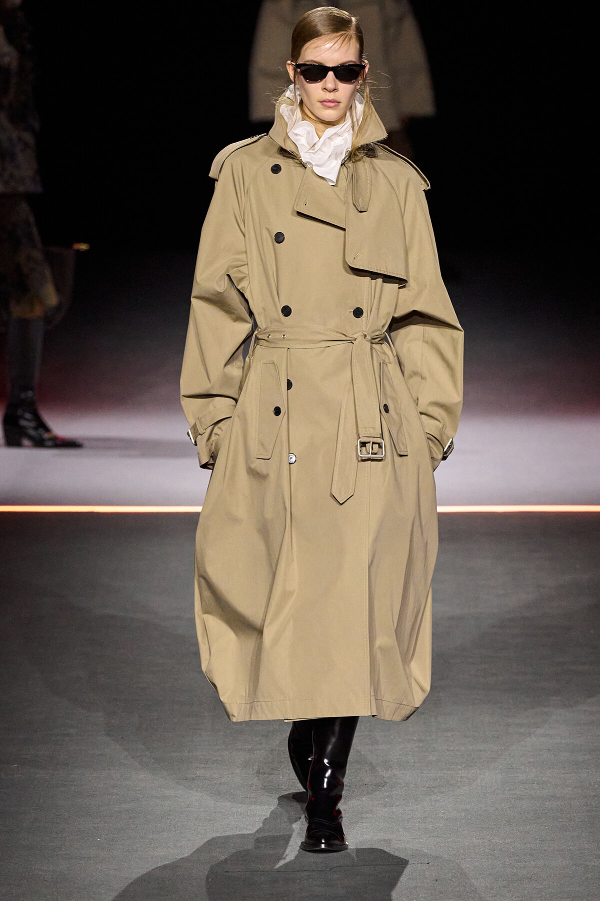 Model walks a runway wearing a knee‑length beige trench coat with a belt, dark sunglasses, and black boots.