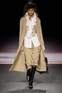 Runway model in a beige trench coat over a white ruffled blouse, corset, beige cargo pants, and black knee-high boots with a black beret and veil.