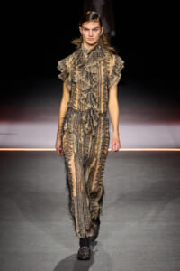 Model on a runway wearing a beige-brown lace-patterned outfit with large ruffled sleeves and matching pants.