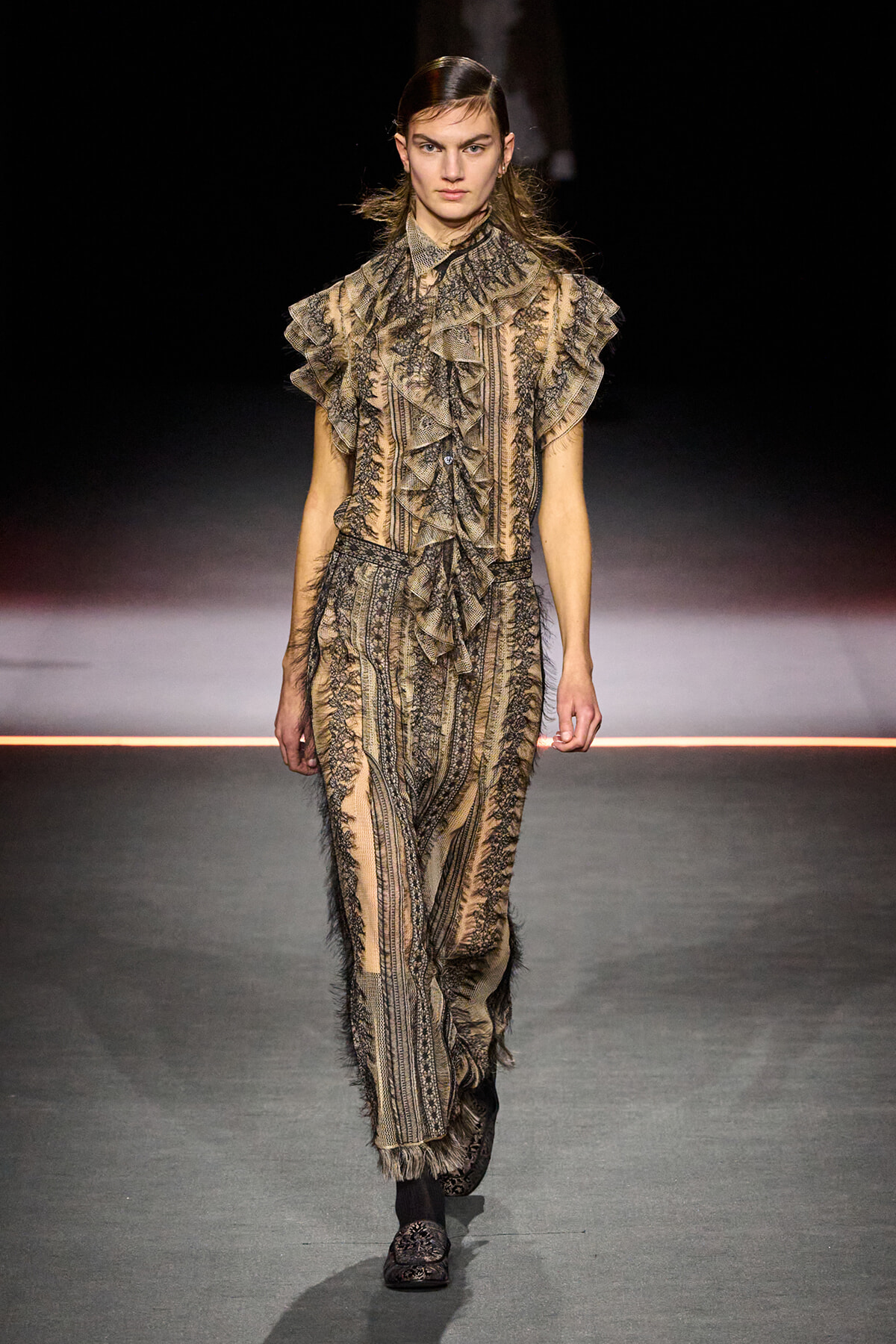 Model on a runway wearing a beige-brown lace-patterned outfit with large ruffled sleeves and matching pants.