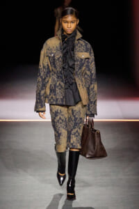 Model walks the runway in a brown and black damask-pattern suit with oversized jacket, black ruffled top, knee-length pants, tall black boots, and a dark brown handbag.