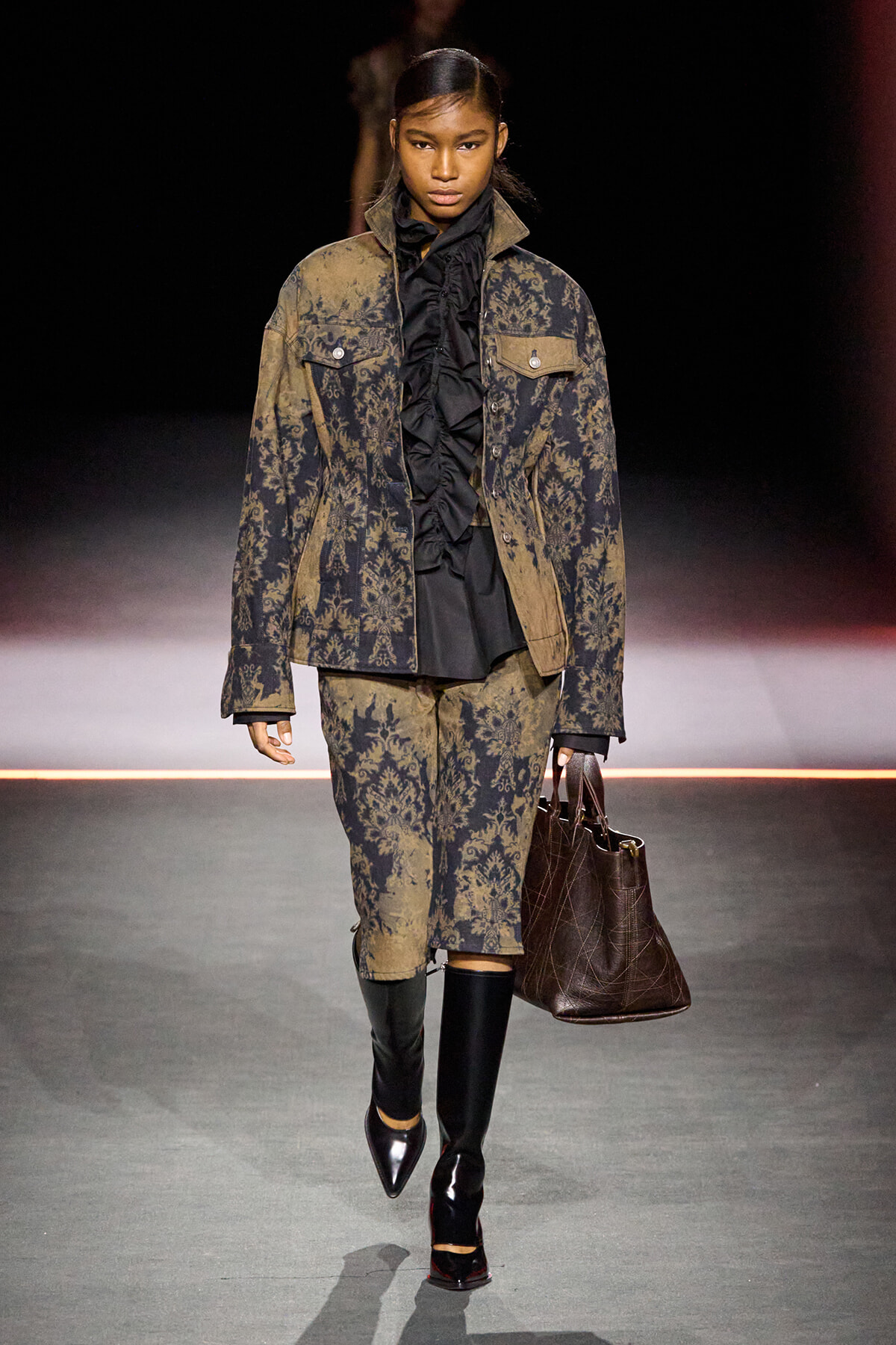 Model walks the runway in a brown and black damask-pattern suit with oversized jacket, black ruffled top, knee-length pants, tall black boots, and a dark brown handbag.