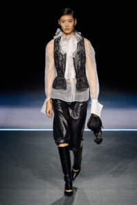 Model walking a runway in a sheer white blouse with a black lace vest over it, black leather shorts, and knee-high boots, holding a black glove accessory.