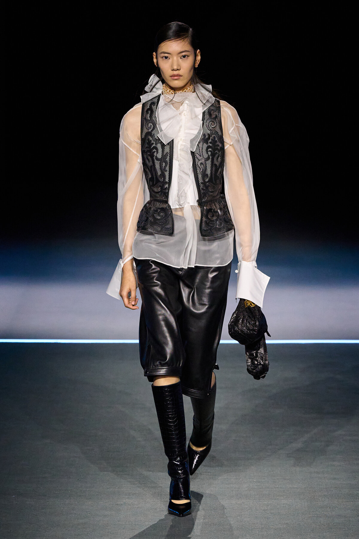 Model walking a runway in a sheer white blouse with a black lace vest over it, black leather shorts, and knee-high boots, holding a black glove accessory.