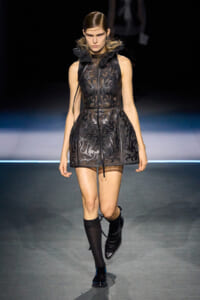 Model walks the runway in a black lace mini dress with a high ruffled collar and sheer panels, paired with black knee-high socks and shoes.