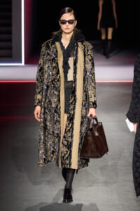 Model walks the runway in a long black-and-gold floral coat with beige panels, black outfit, sunglasses, and a dark brown handbag.